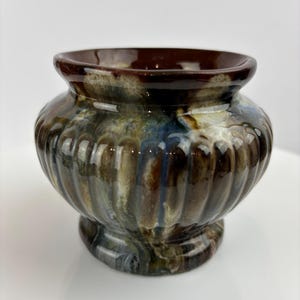 May include: A decorative ceramic vase with a pedestal base. The vase features a ribbed design and a glossy finish with a blend of brown, blue, and green hues. The rim is a darker shade of brown.