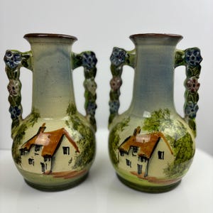May include: Two small ceramic vases with floral handles. Each vase is painted with a cottage scene and green foliage.