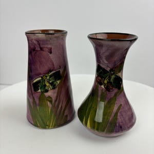 Peut inclure: Deux petits vases en céramique avec un motif floral violet et vert. Chaque vase présente un papillon aux ailes vertes et au corps brun.