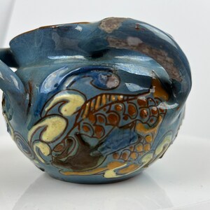 Könnte beinhalten: Ein kleiner blauer Keramiktopf mit zwei Henkeln und einem dekorativen Design aus stilisierten Fischen in Braun, Gelb und Weiß.