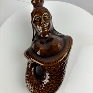 May include: A brown ceramic bottle shaped like a mermaid with a detailed face and a scaly tail.
