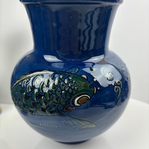 May include: A blue ceramic vase with a raised design of a green and white koi fish. The vase has a round body and a narrow neck.