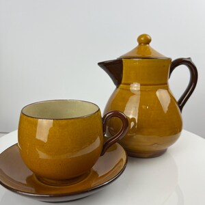 May include: A brown and tan ceramic tea set with a teapot, cup, and saucer. The teapot has a rounded body and a lid with a knob. The cup is round and has a handle. The saucer is round and has a lip.