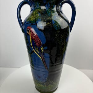 May include: A blue ceramic vase with two handles and a painted bird design. The vase has a dark blue background with green and yellow accents. The bird is red, yellow, and blue.