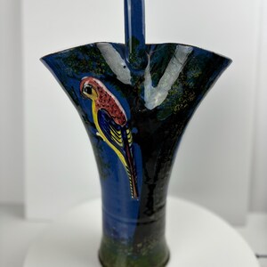 May include: A tall, blue ceramic vase with a handle, featuring a hand-painted parrot design. The vase has a flared top and a dark blue exterior with a painted forest scene. The parrot is red, yellow, and blue.