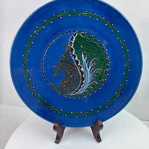 May include: A round ceramic plate with a vibrant blue glaze featuring a central brown and green stylised animal illustration. The design is encircled by a green wavy line with white dots, set against a solid blue background on the plate surface.
