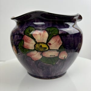 Puede incluir: Un pequeño jarrón de cerámica morado con un borde ondulado. El jarrón está decorado con flores rosas y hojas verdes pintadas a mano.