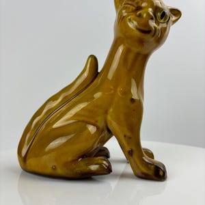 May include: A ceramic figurine of a brown cat with a winking eye and a playful expression. The cat is sitting with its tail curled up behind it.