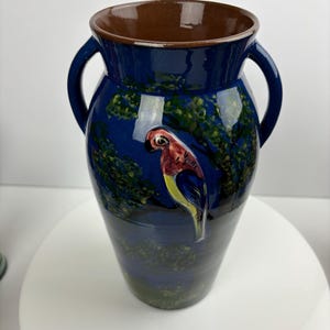 Può includere: Un vaso in ceramica blu con due manici e un pappagallo dipinto sul lato. Il vaso ha un bordo marrone e accenti verdi e gialli.