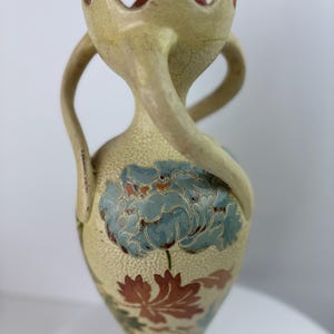 Puede incluir: Un jarrón de cerámica alto, color crema, con superficie texturizada y dos asas curvas. El jarrón presenta un diseño floral azul y detalles de hojas rojas y verdes. La parte superior tiene un borde festoneado único.