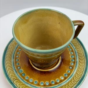 May include: A ceramic teacup and saucer set featuring a warm mustard yellow interior and a deep brown center. The saucer is decorated with a repeating embossed circular pattern and a teal blue rim that matches the teacup's upper edge.