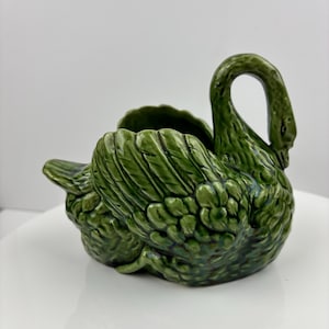 May include: A green ceramic swan planter with detailed feather textures. The swan has a curved neck and a small opening for planting. The planter is a decorative item, suitable for indoor use. The swan is a vibrant shade of green.