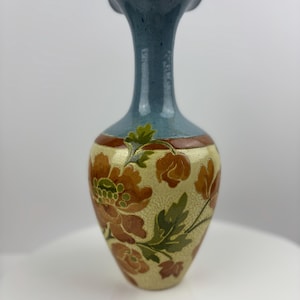 Puede incluir: Un jarrón de cerámica con un borde acampanado y con volantes y un cuello delgado. La parte superior es azul, que pasa a un cuerpo beige adornado con patrones florales marrones y hojas verdes. Una banda marrón rodea el jarrón cerca del cuello.