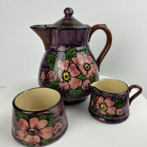 May include: A purple ceramic teapot and two matching creamer and sugar bowls with a floral design. The teapot has a lid and a handle. The creamer and sugar bowls have handles. The flowers are pink with yellow centres and green leaves.