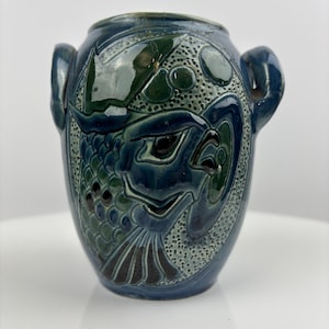 May include: A blue ceramic vase with a fish design. The vase has two handles and is decorated with a detailed relief of a fish, with green and black accents. The vase is approximately 6 inches tall.