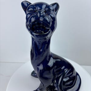 May include: A glossy, navy blue ceramic dog figurine. The dog is sitting upright with its head tilted back, mouth open, and ears perked. The figurine has a smooth, reflective surface and is set against a white background.