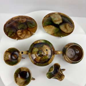 May include: A ceramic tea set with a brown, green, and gold glaze. The set includes a teapot, two cups, a creamer, a sugar bowl, and two saucers. The tea set is arranged on a white surface.