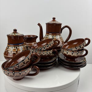 Puede incluir: Un juego de té de cerámica marrón y blanco con una tetera, jarra de crema, azucarero y cuatro tazas y platillos. El juego presenta un patrón geométrico con acentos marrones, blancos y azules.