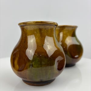 May include: Two small, handcrafted ceramic vases with a glossy, brown glaze. The vases have a rounded body and a slightly flared rim. The glaze has a mottled appearance with hints of green and brown.