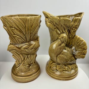 May include: Two ceramic vases with a brown glaze. One vase features a bird with a raised tail, the other features a turkey with a raised tail. Both vases have a textured surface.