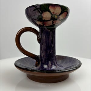 Peut inclure: Un bougeoir en céramique violet avec une poignée brune et un motif floral. Le bougeoir a un petit plat au fond.