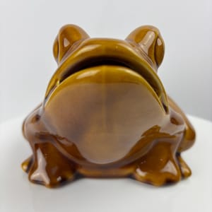 Könnte beinhalten: Eine Froschfigur aus Keramik mit glänzender brauner Glasur. Der Frosch hat große Augen und einen offenen Mund. Die Figur steht auf einer weißen Oberfläche. Dekoratives Objekt.