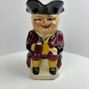 Könnte beinhalten: Keramikfigur eines Mannes mit Dreispitz und bordeauxrotem Mantel, der einen Krug hält. Die Figur hat ein lächelndes Gesicht, gelbe Kniebundhosen und schwarze Schuhe. Die Figur sitzt auf einem kleinen, rechteckigen Sockel.