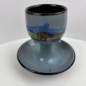 Może przedstawiać: Ceramiczny kubek na jajko z podstawą w kształcie spodka. Kubek jest w stonowanym niebieskim kolorze z ciemną obwódką i namalowaną sceną krajobrazu z brązowym koniem. Spodek i nóżka również są niebieskie.