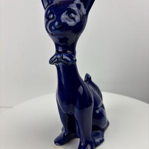 May include: A glossy, navy blue ceramic cat figurine. The cat is sitting upright with its ears perked up and a small bow tie around its neck. The figurine has a smooth, reflective surface and is set against a white background.