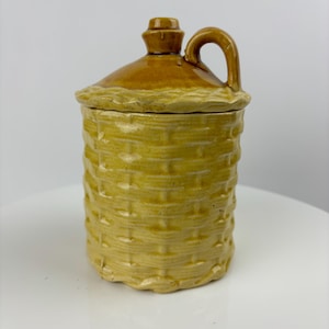 May include: A yellow ceramic jar with a woven pattern and a lid. The jar has a handle and a spout.