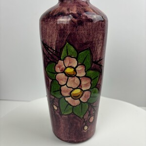 Peut inclure: Un vase cylindrique et élancé avec un fond violet foncé et un motif floral. Les fleurs sont roses avec des centres jaunes et des feuilles vertes. Le vase mesure environ 20 cm de haut.