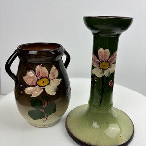 Pode incluir: Um conjunto de dois vasos de cerâmica vintage com desenhos florais. O vaso menor tem duas alças e um esmalte marrom com folhas verdes e uma flor rosa. O vaso mais alto é um castiçal com um esmalte verde e uma flor rosa.