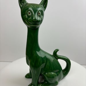 May include: A green ceramic cat figurine with a long neck and a wide smile. The cat has large, round eyes with gold pupils.