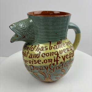 May include: A green and yellow ceramic pitcher with a fish head spout and a handle. The pitcher is decorated with a brown inscription that reads "The world has battle fought and conquer ever rise. Or if ye fall Pray God a gen"