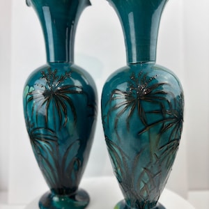 Op de afbeelding: Twee turquoise keramische vazen met een glanzende afwerking. Elke vaas heeft een uitlopende, gerimpelde bovenkant en een bolvormig lichaam met een zwart bloemmotief. De vazen hebben een voet en zijn ongeveer 30 cm hoog.