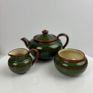 Puede incluir: Un juego de té de cerámica verde y marrón con tetera, jarra de crema y azucarero. El juego tiene un diseño sencillo con ribete marrón.