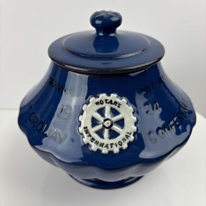 Puede incluir: Un tarro de cerámica azul oscuro con tapa y pomo. El tarro tiene un emblema Rotary International blanco y plateado y el texto "Torquay 19 Conference". El tarro tiene un diseño ondulado alrededor de la base.