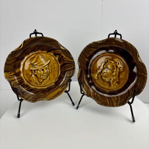 May include: Two brown ceramic plates with raised images of people's faces. The plates have a woodgrain pattern and are on black metal stands.