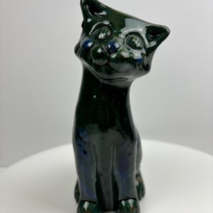 May include: A green ceramic cat figurine with blue eyes and brown accents. The cat is sitting upright with its head tilted up.