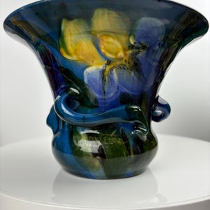 May include: A blue ceramic vase with a wide mouth and a floral design. The vase has a unique, twisted handle and is decorated with yellow and purple flowers.