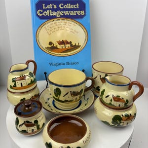 Könnte beinhalten: Ein Vintage-Set aus Cottageware-Keramik in Creme, Braun und Grün mit handbemalten Szenen von Häusern und Bäumen. Das Set beinhaltet eine Teekanne, eine Milchkännchen, eine Zuckerdose, eine Tasse und eine Untertasse sowie eine kleine Schale. Das Buch "Let's Collect Cottagewares" von Virginia Brisco ist ebenfalls abgebildet.