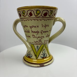 May include: A vintage ceramic cup with two handles, cream-coloured with yellow and brown decorative accents. The cup has a flared base and a rim with a repeating pattern. The text on the cup reads: "you your lips would keep from five things obs with care."