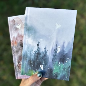 Könnte beinhalten: Zwei Notizbücher mit Aquarellillustrationen einer Waldszene und schwebenden Geistern. Das Cover auf der rechten Seite zeigt einen nebligen blau-grünen Wald mit weißen Geistern. Das linke Cover zeigt eine rotbraune Waldszene mit einem weißen Geist.