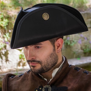 May include: A black tricorn hat with a gold button on the top. The hat is made of leather and has a wide brim. The hat is being worn by a person with a beard.