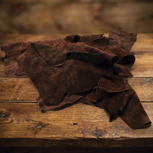 Puede incluir: Un montón de retales de cuero marrón oscuro con un aspecto texturizado, similar al ante. El cuero está dispuesto sobre una superficie de madera rústica, con vetas y nudos visibles. La iluminación crea sombras y resalta la textura del cuero.