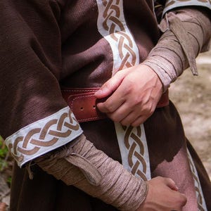 May include: A brown tunic with white and brown braided trim. The tunic has a brown leather belt with a metal buckle. The sleeves are rolled up and secured with a brown fabric tie.
