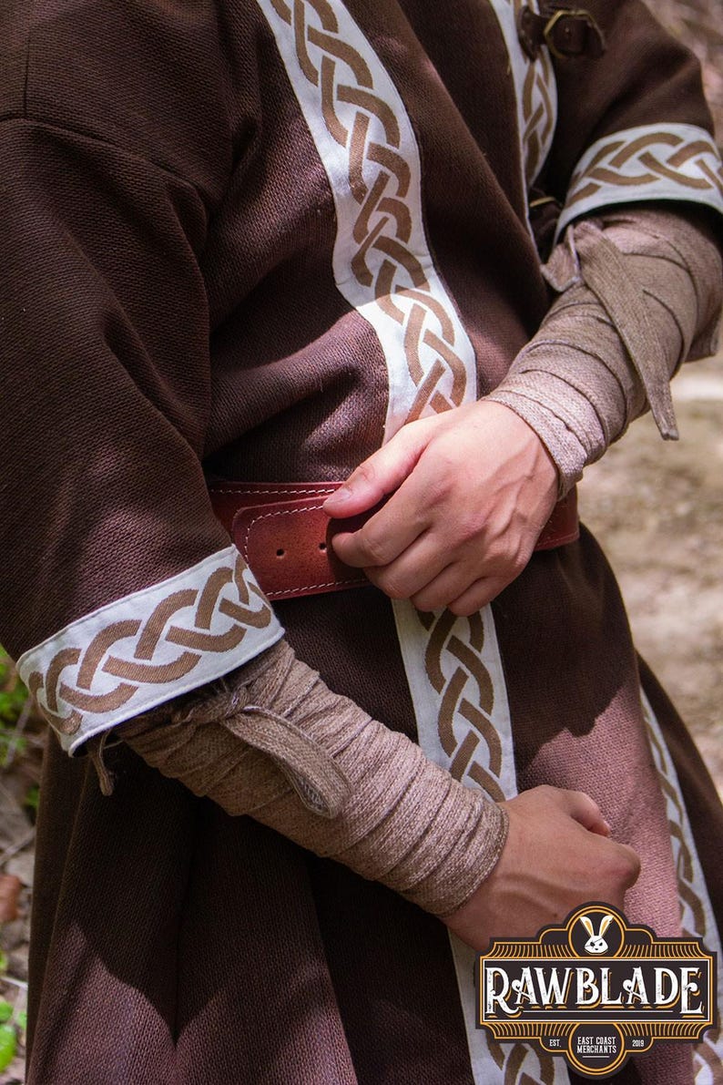May include: A brown tunic with white and brown braided trim. The tunic has a brown leather belt with a metal buckle. The sleeves are rolled up and secured with a brown fabric tie.
