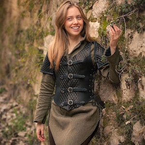 May include: A woman wearing a black leather vest with a quilted design and buckles. The vest has a laced-up back and is worn over a green shirt. The woman is standing in front of a stone wall with vines growing on it. The image includes the logo "Rawblade East Coast Merchants Est. 2018."