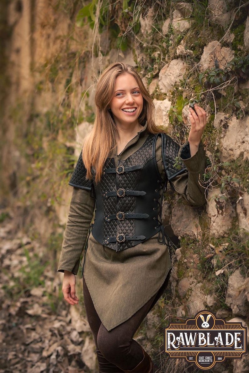 May include: A woman wearing a black leather vest with a quilted design and buckles. The vest has a laced-up back and is worn over a green shirt. The woman is standing in front of a stone wall with vines growing on it. The image includes the logo "Rawblade East Coast Merchants Est. 2018."