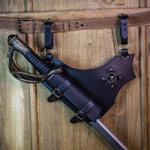 Könnte beinhalten: Ein schwarzes Lederschwert-Scheide mit einer dekorativen Metallschnalle und einem Riemen zum Befestigen an einem Gürtel. Die Scheide hängt an einer Holzwand mit einem braunen Ledergürtel.